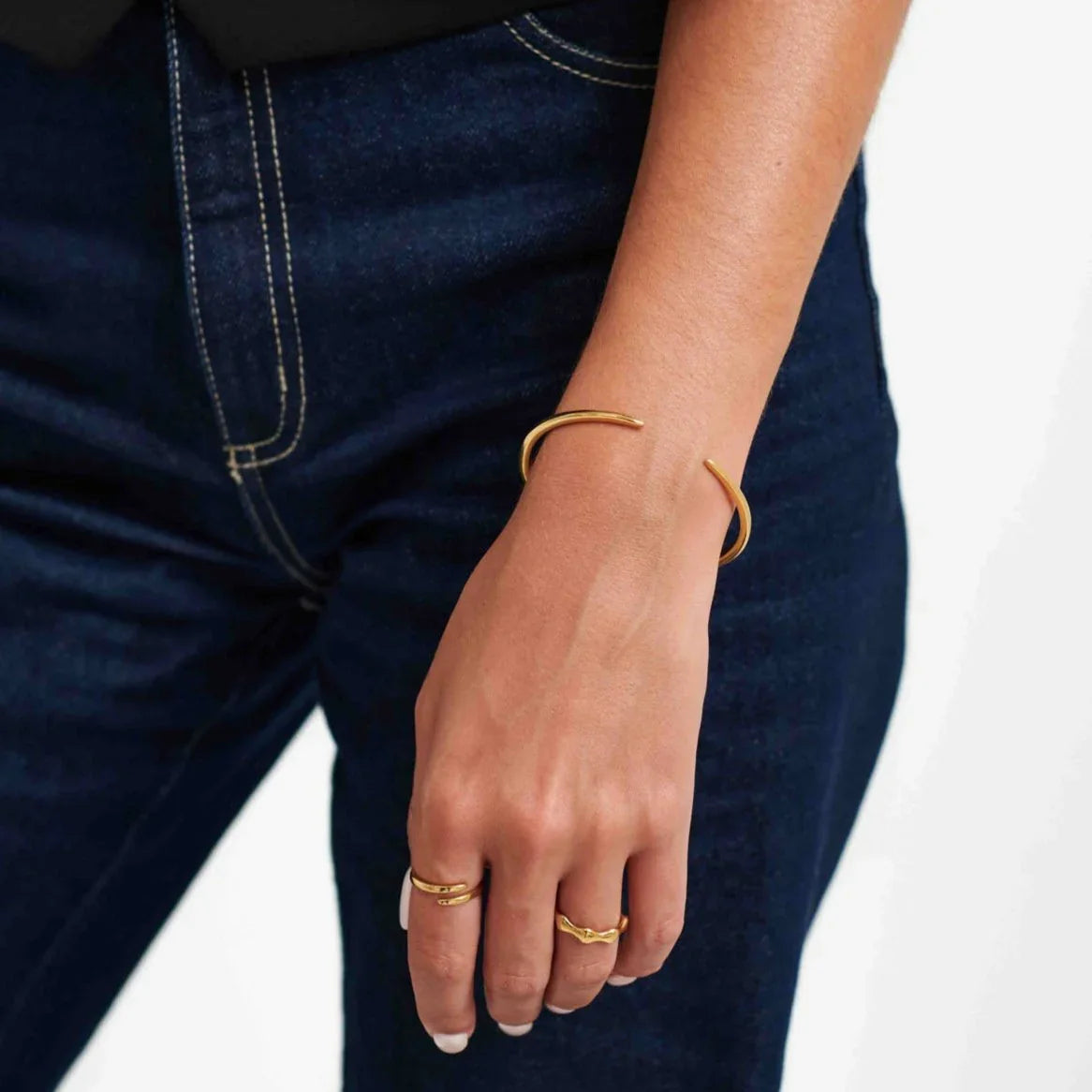 Close-up of hand wearing minimalist gold rings and cuff bracelet with dark blue jeans background