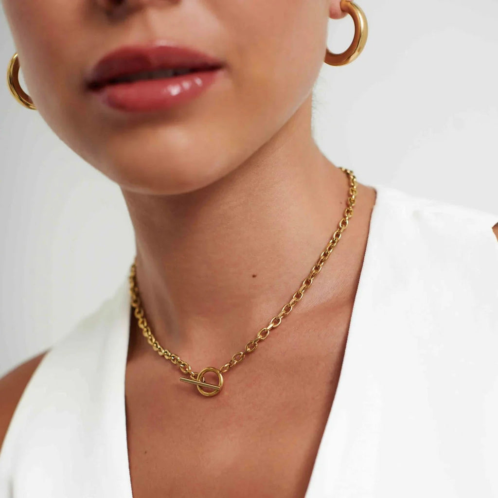 Close-up of woman wearing gold hoop earrings and chain necklace with toggle clasp on white top