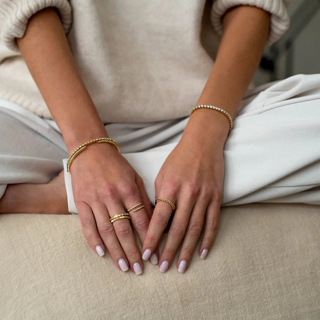 Close-up of hands wearing gold rings and bracelets on a beige fabric surface, dressed in light beige and white clothes