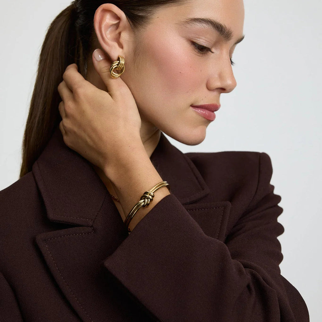 Model wearing gold knot design hoop earrings and matching gold knot bangle bracelet with dark brown blazer
