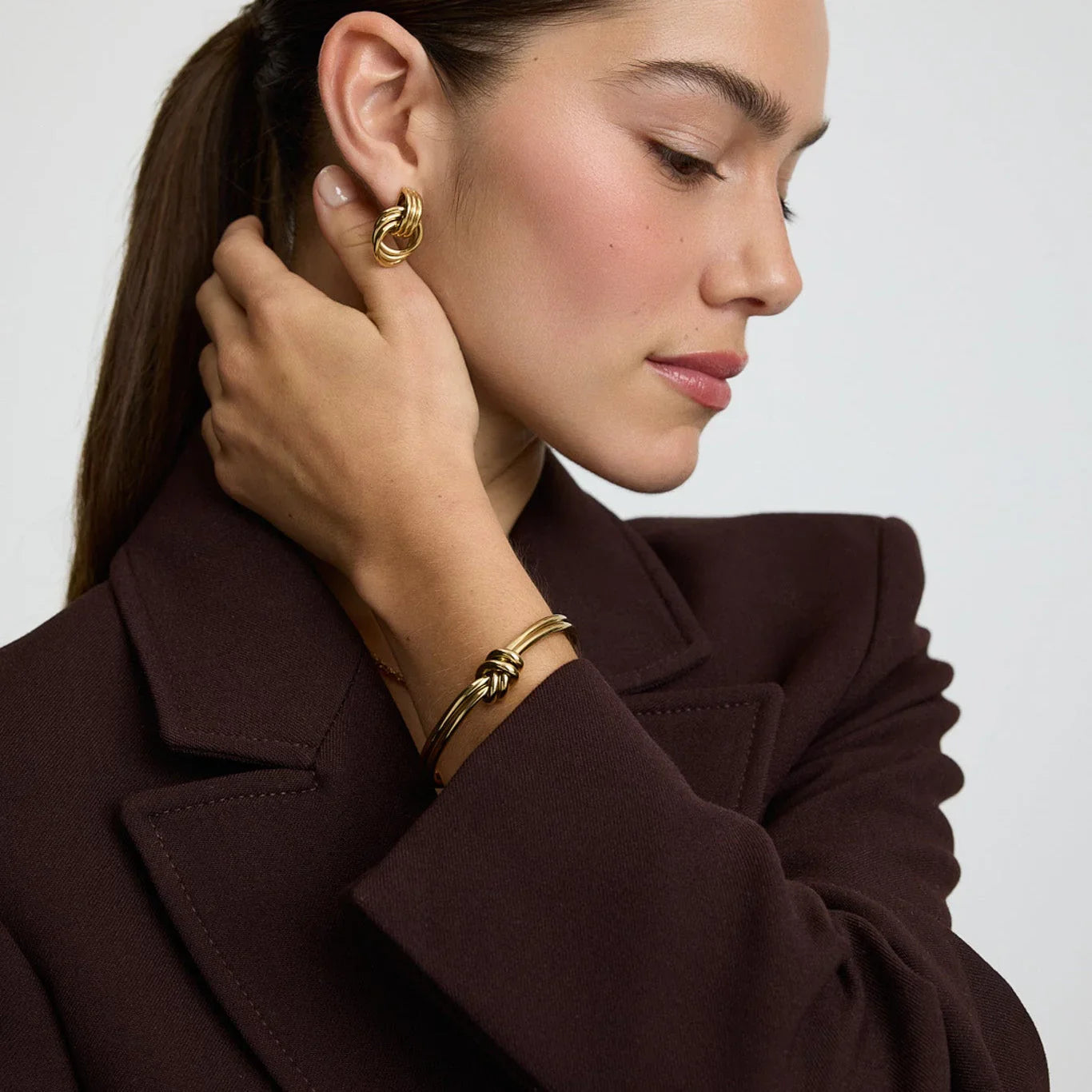Model wearing gold knot design hoop earrings and matching gold knot bangle bracelet with dark brown blazer
