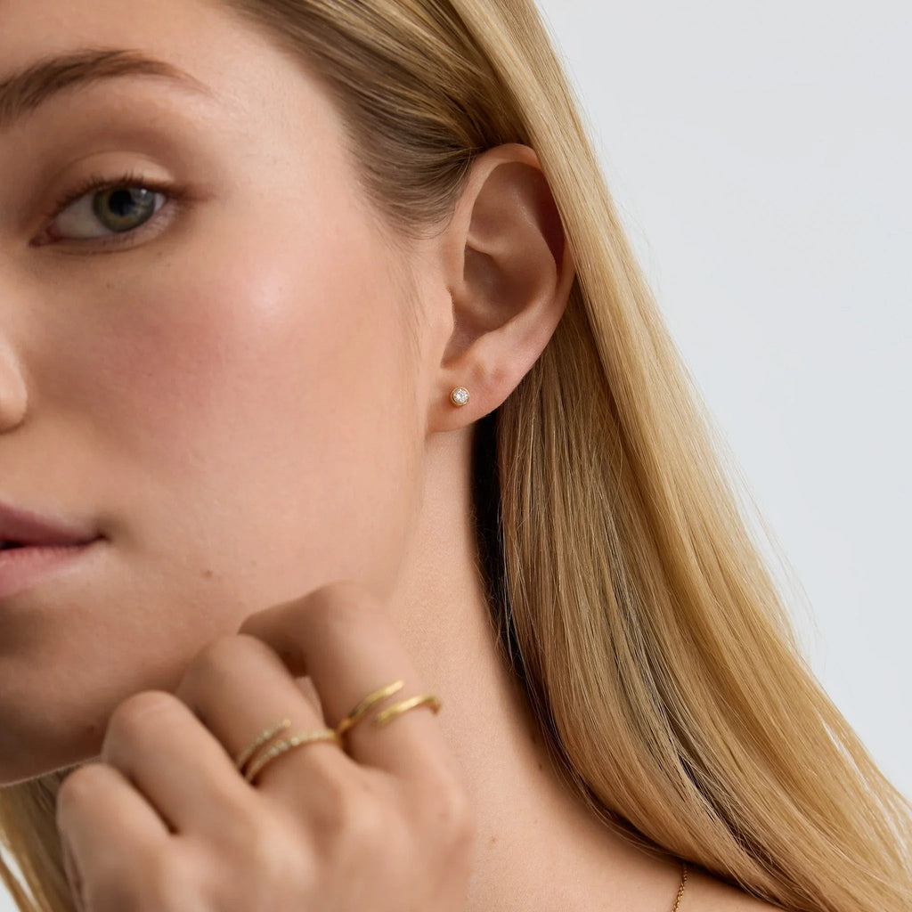Close-up of woman wearing small diamond bezel stud earring and gold rings on hand